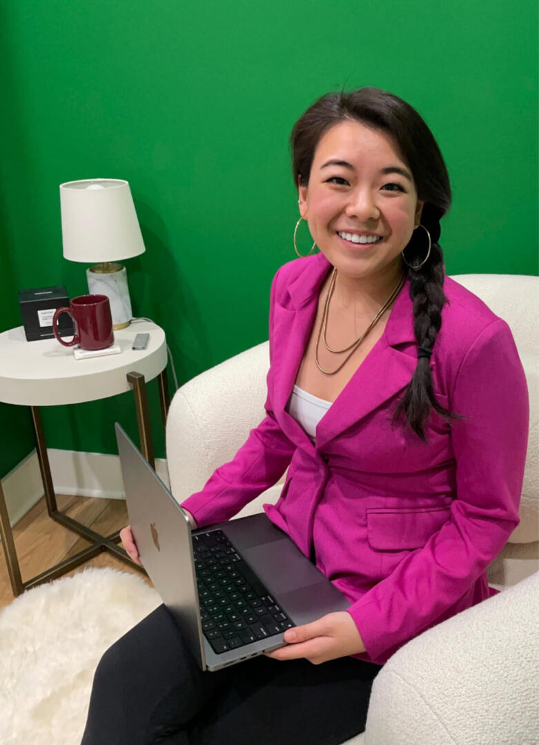 Vivian, an East Asian woman in a hot pink blazer, smiles and sits on a beige armchair with a laptop open in her lap